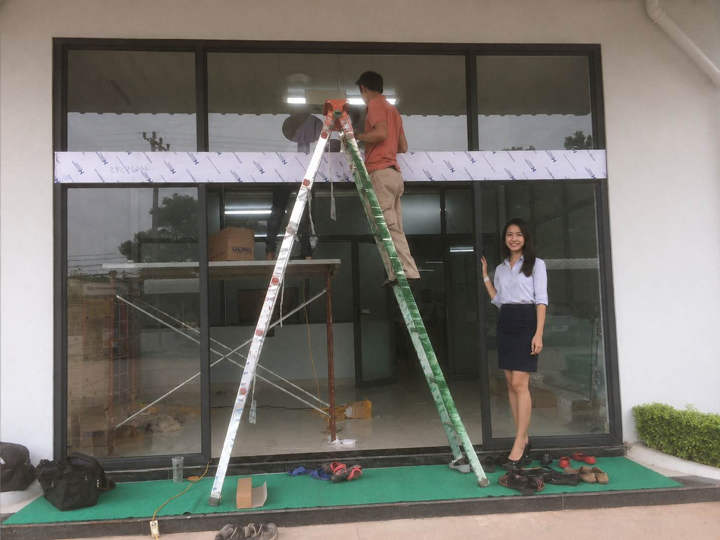 Thi công lắp đặt cửa kính mặt tiền văn phòng với thợ đứng trên thang và nhân viên nữ đứng cạnh